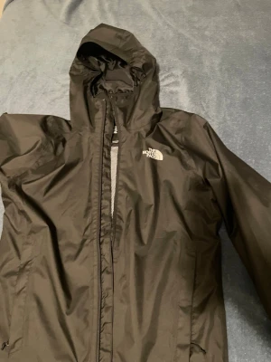 Svart regnjacka från The North Face - Snygg svart regnjacka från The North Face med huva och dragkedja framtill. Jackan har logga på bröstet och ryggen, samt är tillverkad i ett vattenavvisande material. Perfekt för regniga dagar och outdoor-äventyr.