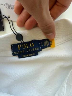 Vit pikétröja från Polo Ralph Lauren - Klassisk vit pikétröja från Polo Ralph Lauren. Tröjan har krage, korta ärmar och knappar vid halsen. Modellen är custom slim fit och tillverkad i bomull för en skön känsla. ALDRIG ANVÄND. Säljer för jag fick den i present och har en likadan! Pris 699kr