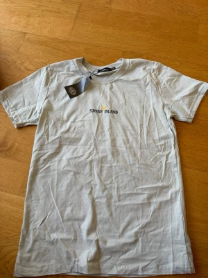 Vit/beige Stone Island t-shirt i bomull - Snygg vit t-shirt från Stone Island med klassisk logga broderad i svart och gult på bröstet. T-shirten är kortärmad och har en rund halsringning. Tillverkad i mjuk bomull för en skön känsla och clean look.