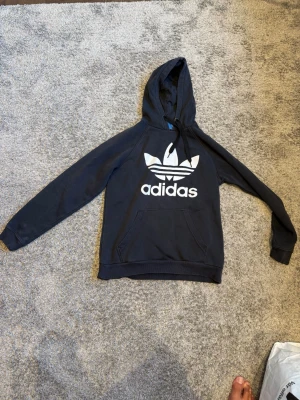 Svart Adidas hoodie med logga - Snygg svart hoodie från Adidas med stor vit Trefoil-logga och text på bröstet. Klassisk känguruficka framtill och huva med dragsko. Tillverkad i mjukt material som passar perfekt till en avslappnad streetwear-look.