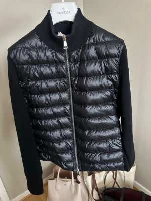 Svart cardiganjacka från Moncler - Snygg svart cardiganjacka från Moncler med quiltad, glansig framsida och stickade ärmar. Jackan har hög krage, dragkedja framtill och Moncler-logga på ärmen. Använd ett par fåtal gånger. Kvitto finns och Moncler galge med kommer. Storlek M men passar även bra som S 