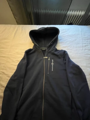 Marinblå hoodie från Sail Racing - Snygg marinblå hoodie från Sail Racing med dragkedja framtill och logga på bröstet. Tröjan har huva, lång ärm och en extra bröstficka med dragkedja. Perfekt för dig som gillar sportig och stilren look.
