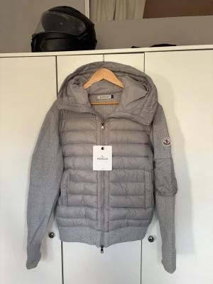Grå dunjacka med stickade ärmar Moncler - Snygg grå dunjacka från Moncler med stickade ärmar och huva. Jackan har quiltad front, två sidofickor med dragkedja och ribbade muddar. Moncler-logga på vänster ärm. Perfekt för kalla vinterdagar och riktigt trendig look.