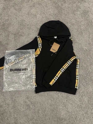 Svart Burberry hoodie med rutiga detaljer - Säljer en svart hoodie från Burberry med klassiska beige, röda och svarta rutiga detaljer längs ärmarna och axlarna. Hoodien har dragsko i huvan och en stor ficka framtill. Perfekt för dig som vill ha en stilren men ändå lyxig look.