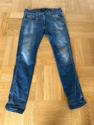 Blå Replay jeans med slitningar - Säljer ett par blå jeans från Replay med snygga slitningar och tvättade detaljer. Klassisk femficksmodell med raka ben och normal midja. Jeansen har en modern look och är tillverkade i stretchigt denimtyg för extra komfort.