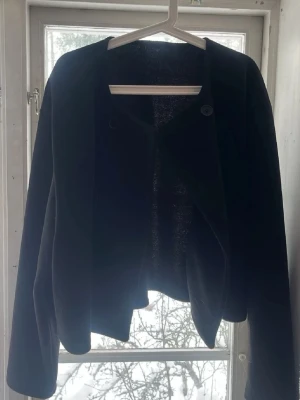 Kappa - Stilren svart kappa med dubbelknäppning framtill och rak passform. Kappan är kortare i modellen och har en enkel, kraglös design. Perfekt för dig som gillar minimalistisk stil och vill ha en klassisk look. Materialet ser ut att vara ullblandning. Pefekt att ha nu till höst!🍁🍂