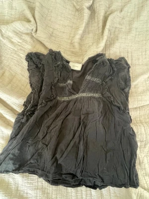Svart blus med volang från Neo Noir - Supersöt svart blus från Neo Noir med volangdetaljer och spets längs bröstet och axlarna. Blusen har v-ringning och är ärmlös. Materialet känns lätt och luftigt. Solblekt.