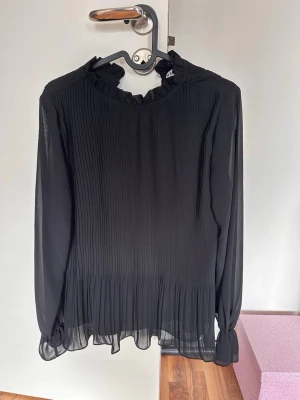Svart plisserad blus med volangkrage - Snygg svart blus med plisserat tyg och volangdetalj vid halsen. Blusen har långa, transparenta ärmar och en lös passform. Tillverkad i ett lätt och luftigt material som ger en elegant look.