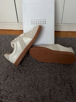 Beige vita masion marigela skor  - Snygga beige masion marigela skor med detaljer i mocka och slät textil. Skorna har en klassisk låg profil och bruna gummisulor med bra grepp. Perfekta för dig som gillar stilrena och tidlösa sneakers med neutrala färger.