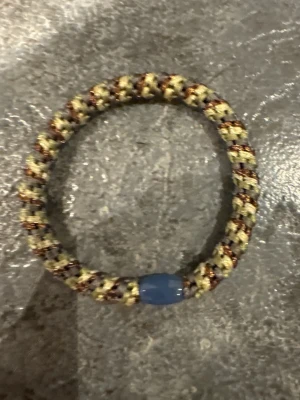 Flätat armband med blå pärla - Snyggt armband med flätad design i beige, brunt och svart. Armbandet har en blå pärla som detalj och är gjort av slitstarkt snöre. Perfekt accessoar för en cool och avslappnad stil.