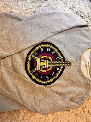 Grå Kenzo Paris sweatshirt med tryck - Snygg grå sweatshirt från Kenzo med stort broderat tryck på bröstet föreställande Eiffeltornet och texten 'Kenzo Paris' i gult, svart och lila. Tröjan har rund hals och är tillverkad i mjukt material, perfekt för en trendig streetstyle-look.