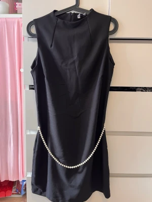 Svart kort klänning med pärldetalj - Snygg svart ärmlös kort klänning med hög hals och dekorativ pärlkedja längs höften. Klänningen har en enkel, elegant design och dragkedja i ryggen. Perfekt för dig som gillar stilrena plagg med en twist.