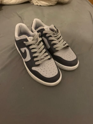 Nike Dunk Low grå och marinblå - Snygga Nike Dunk Low sneakers i grått och marinblått läder med klassisk vit sula och matchande grå skosnören. Ikonisk Swoosh på sidan och perforerad tå för extra ventilation. Perfekta för dig som gillar stilrena och sportiga sneakers.