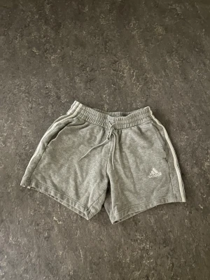 Grå shorts med snörning från Adidas - Säljer ett par grå mjuka shorts från Adidas med vita ränder längs sidorna och snörning i midjan. Shortsen har elastisk midja och två sidofickor. Perfekta för chill dagar eller träning.