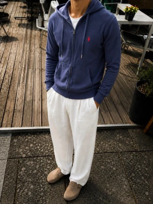 Polo Ralph Lauren Marinblå Zip Hoodie – Storlek M - Snygg och stilren marinblå zip-hoodie från Polo Ralph Lauren. Klassisk modell med den ikoniska röda Polo Pony-loggan broderad på bröstet.  Hoodien har dragkedja framtill, huva med justerbara snören, känguruficka och ribbade muddar vid ärmar och nederkant. Tillverkad i 100 % bomull. En tidlös hoodie som passar perfekt till jeans, chinos eller en mer avslappnad outfit med vita byxor.  • Märke: Polo Ralph Lauren • Färg: Marinblå • Storlek: M • Material: 100 % bomull