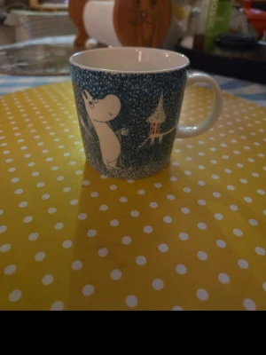 Muminmugg " Lätt snöfall" - Säljer en blåvit muminmugg med vintermotiv och snöfall. Muggen har ett tryck av Mumin och Muminhuset, och är tillverkad i porslin. Perfekt för te eller kaffe och har ett bekvämt handtag.