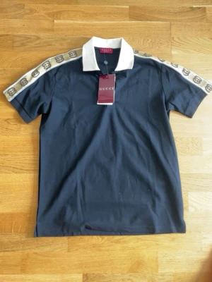 Svart pikétröja med logga - Snygg svart pikétröja med vit krage och beige band med GG-logga på axlarna. Klassisk passform och korta ärmar, perfekt för en trendig och avslappnad look.