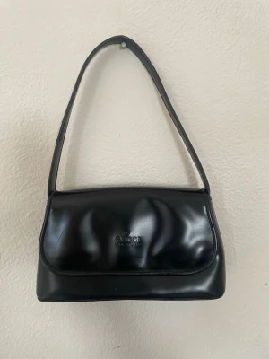 Vintage väska💕 - Stilren svart axelremsväska  i slätt läder. Väskan har en klassisk rektangulär form med lock och präglad logotyp på framsidan. Enkel design som ger en tidlös och elegant look. Perfekt för dig som gillar lyxiga accessoarer. Köpt sacond hand tror ej den är äkta men världens snyggaste väska😁