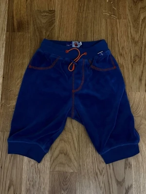 Blå / orange barn / baby byxor från Molo - Storlek 62 - Ett par fina blå babybyxor från Molo med snygga orangea detaljer! Byxorna har resår i midjan och muddar vid bensluten för en bekväm passform. Midjan kan enkelt justeras med ett orange dragsnöre. På baksidan finns ett dekorativt orange sicksackmönster som ger byxorna en extra fin design! Storlek: 62. Material: 75 % Bomull och 25 % Polyester. Säljs i bra skick! Kika gärna in på mina andra annonser, jag säljer just nu flera barnplagg! Hör av dig vid frågor och intresse!