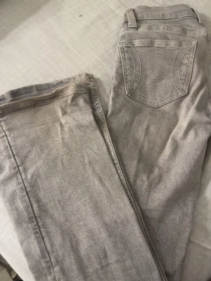 Ljusgrå bootcut jeans från Gina - Säljer ett par ljusgrå jeans från Gina i bootcut modell. Jeansen har klassiska fickor bak med dekorativa sömmar och bälteshällor. Materialet är mjukt denim som passar perfekt till en avslappnad stil.