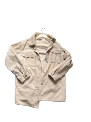 Beige overshirt i manchester - Snygg beige overshirt i manchestertyg med klassisk krage och två stora bröstfickor. Knäpps med silverfärgade knappar framtill och har långa ärmar med knapp vid ärmslut. Perfekt lager på lager-plagg för en avslappnad stil.