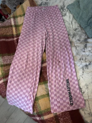 Lila rutiga pyjamasbyxor SMILEYWORLD - Lila och vita pyjamasbyxor med rutigt mönster och trycket 'SMILEYWORLD' på ena benet. Byxorna har resår i midjan och är tillverkade i mjukt bomullstyg. Perfekta för chill hemma eller mysiga kvällar. Hårfärgs fläck på ena benet som inte går bort i tvätten 