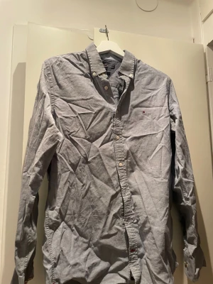 Ljusblå skjorta från Tommy Hilfiger - Snygg ljusgrå skjorta från Tommy Hilfiger i slim fit-modell med stretch. Skjortan har klassisk krage, knappar framtill och en liten broderad logga på bröstet. Perfekt för en stilren look.