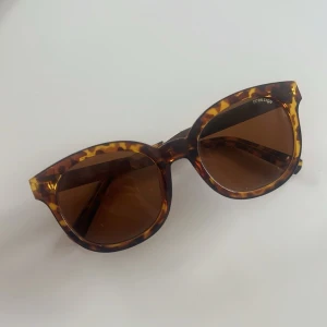 Bruna solglasögon i leopardmönster - Snygga solglasögon med rundad form och leopardmönstrad båge i bruna toner. Glasen är mörkbruna och ger en cool retrovibe. Perfekta för soliga dagar när du vill sticka ut lite extra.