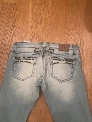 Lågmidjade vintage jeans - Lågmidjade vintage jeans med revor. De har fastsydda detaljer och en cool ”wash” Midjemåttet är 41cm, innerbenslängden är 84cm och måttet från gren till midja är ca 19cm.