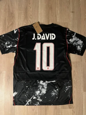 Svart Kanada fotbollströja David #10 - Snygg svart fotbollströja med Kanadas landslagsmärke och nummer 10 på bröstet. Tröjan har vitt splattermönster, röda detaljer och J. David tryckt på ryggen. Tillverkad i lätt och ventilerande material, perfekt för träning eller match. HELT NY!