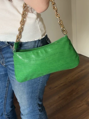 Grön axelväska med kedjerem - Snygg grön handväska i croco-mönstrat konstläder med guldfärgad kedjerem. Väskan har en avlång form och stängs med dragkedja upptill. Perfekt accessoar för att lyfta din outfit med en poppig färg och trendig design.