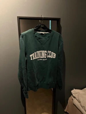 Grön sweatshirt från Björn Borg - Mörkgrön sweatshirt från Björn Borg med vit text 'TRAINING CLUB' på bröstet. Klassisk rund halsringning och ribbade muddar vid ärmslut och nederkant. Perfekt för en sportig och avslappnad stil.