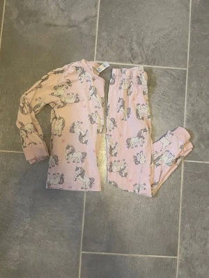 Rosa pyjamas med enhörningsmönster för barn - Söt tvådelad pyjamas för barn i ljusrosa med tryck av enhörningar i vitt, lila och gult. Setet består av långärmad tröja och långa byxor med resår i midjan. Perfekt för mysiga kvällar och sömn. Materialet känns mjukt och bekvämt mot huden.