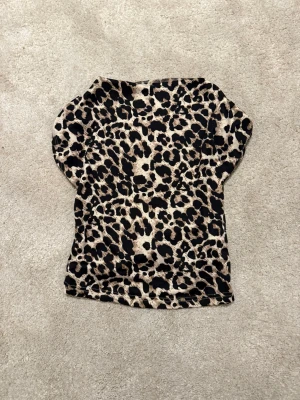 Leopardmönstrad topp med kort ärm - Snygg topp med leopardmönster i beige och svart. Modellen är figurnära med korta ärmar och rund hals. Tillverkad i ett stretchigt syntetmaterial som sitter bekvämt och markerar siluetten. Perfekt för dig som vill sticka ut lite extra. I storlek M men mer som en S!