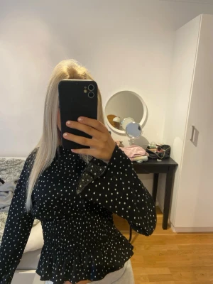 Svart prickig blus med smock - Supersöt svart blus med vita prickar och smock i midjan som ger en snygg siluett. Blusen har långa ärmar och volangkant nedtill. Perfekt till jeans eller kjol för en trendig look.