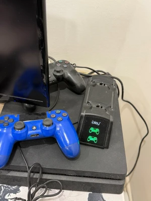 PlayStation 4 med två handkontroller - Säljer en PlayStation 4 konsol i svart med två DualShock 4 handkontroller, en blå och en svart. Medföljer även en laddstation för kontrollerna. Perfekt för gamingkvällar och multiplayer. Klassisk fyrkantig design i plast.