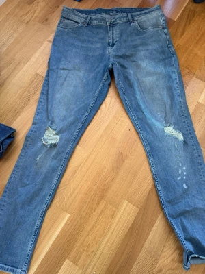Blå slitna jeans med hål - Ett par blå jeans med skinny fit och slitningar på båda knäna. Jeansen har klassisk femficksdesign och är tillverkade i denim. Perfekta för en avslappnad och trendig look.
