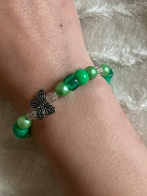 Grönt pärlarmband med fjärilar -  Armband med gröna och genomskinliga pärlor samt två metallfärgade fjärilsberlocker. Armbandet har en mix av glansiga och matta pärlor i olika nyanser av grönt och silverdetaljer. 😊