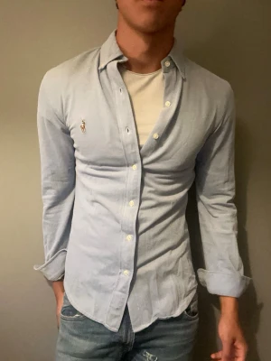 Ljusblå skjorta från Ralph Lauren - Klassisk ljusblå skjorta från Ralph Lauren med broderad logga på bröstet. Skjortan har lång ärm, knappar framtill och är tillverkad i mjuk bomull. Perfekt till jeans eller chinos för en clean och stilren look.