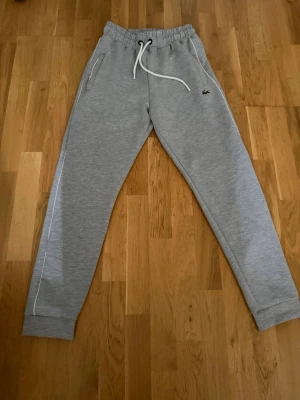 Grå mjukisbyxor från Lacoste - Snygga grå mjukisbyxor från Lacoste med vit dragsko i midjan och klassisk krokodillogga på vänster sida. Byxorna har sidofickor och vita detaljer längs benen. Perfekta för en avslappnad och sportig stil.