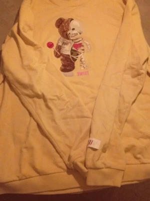 Gul sweatshirt med nalletryck - Cool gul sweatshirt med ett stort tryck av en nalle på framsidan, där ena halvan är skelett och andra halvan är mjukisdjur. Texten 'SWEET' och 'BITTER' syns under motivet. Tröjan har långa ärmar och ribbade muddar. Perfekt för dig som gillar unika prints!