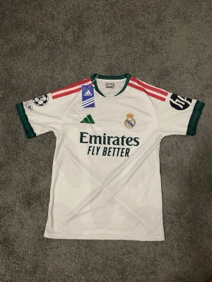 Real Madrid matchtröja Adidas vit ny real Madrid dress till 2027 - Snygg vit Real Madrid matchtröja från Adidas med gröna detaljer och röda ränder på axlarna. Tröjan har klubbmärke, sponsortryck och Champions League-märke på ärmen. Perfekt för fotbollsfans som vill sticka ut på planen eller läktaren.