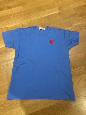 Blå t-shirt från Comme des Garçons - Blå t-shirt från Comme des Garçons PLAY med det ikoniska röda hjärtat broderat på bröstet. Klassisk passform och rund halsringning. Perfekt för dig som gillar stilrena plagg med en twist. Är large men sitter som medium.