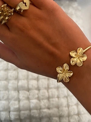 Guldigt armband med blommor - Snyggt armband i guldfärgad rostfritt stål metall med två stora blommor som dekoration. Blommorna har fem blad och ger en cool och trendig look. Perfekt för dig som vill sticka ut med dina accessoarer och gillar detaljer som syns.