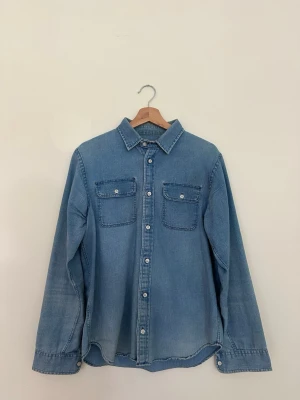 Blå jeansskjorta - Ljusblå jeansskjorta från Real + Simple. Klassisk modell som passar till både vardag och fest. Fint begagnat skick utan anmärkningar. Skickas eller hämtas enligt överenskommelse.