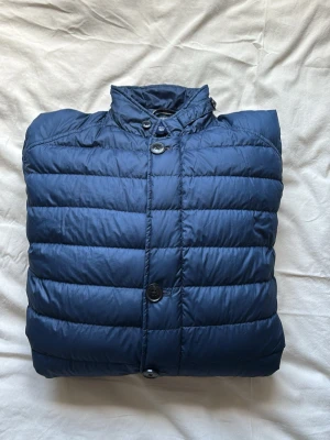 Mörkblå dunjacka från Gant med knappar - Snygg mörkblå dunjacka med quiltad design och raka sömmar. Jackan har ståkrage, knäppning med stora knappar framtill och två fickor med lock. Perfekt för kalla vinterdagar och ger en stilren look.