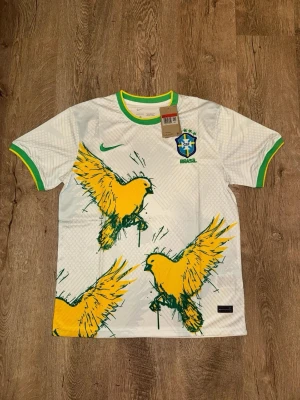 Vit Brasilien fotbollströja med fåglar  - Snygg vit fotbollströja från Nike med Brasilien-emblem på bröstet och gröna, gula detaljer vid krage och ärmslut. Neymar jr nummer 10 på ryggen av tröjan. Tröjan har stora gula papegojor tryckta framtill och ett grönt Nike-märke. Perfekt för dig som älskar fotboll och vill sticka ut på planen. M