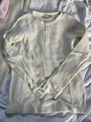 Offwhite långärmad henleytröja - Säljer en offwhite långärmad henleytröja med tre knappar framtill. Tröjan har en ribbad struktur och rundad halsringning. Perfekt för lager på lager och enkel att matcha med jeans eller shorts och är inte använd alls men har ej kvitto eller tag kvar på den.