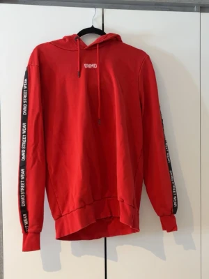 Röd hoodie från DNMD Street Wear - Säljer en röd hoodie från DNMD Street Wear med huva och dragsko. Tryckt logga på bröstet och text längs båda ärmarna. Hoodien har ribbade muddar och en något längre bakdel. Perfekt för en avslappnad streetwear-look.