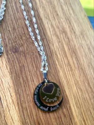 Halsband med berlock ”Love you to the moon and back” - Snyggt halsband i silverfärgad rostfritt stål med ett hänge i form av en måne och ett hjärta. På månen står det 'to the moon and back' och på hjärtat 'I love you'. Perfekt för dig som vill bära något personligt och betydelsefullt. Kedjan är ca 50 cm lång 
